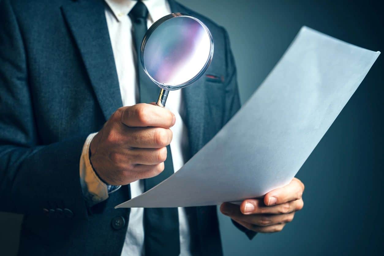 A man in a business suit holding a magnifying glass to a sheet of paper.